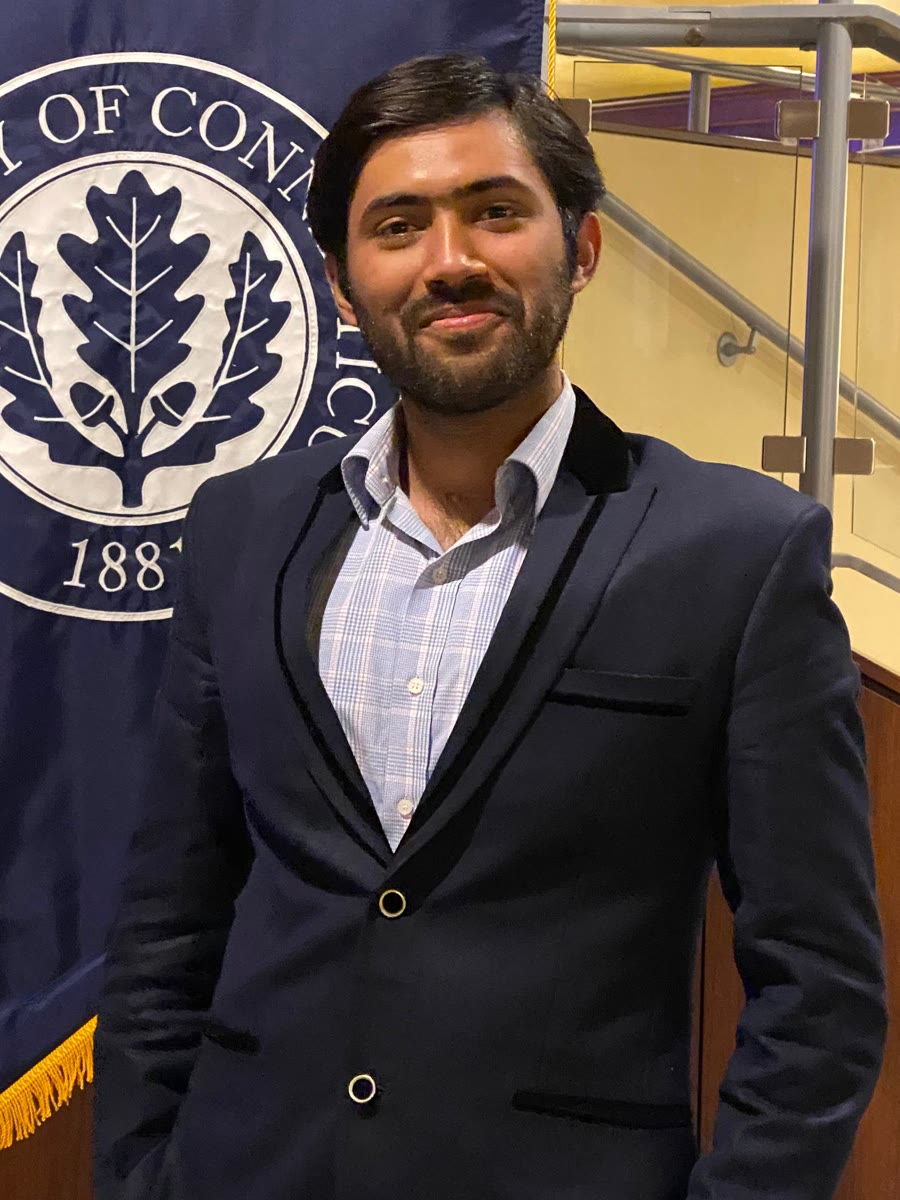 Musawir Abrar in a dark blazer, smiling slightly, in front of a University of Connecticut banner.