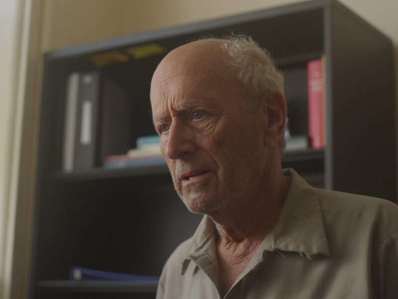 An older man in a frayed khaki work shirt, mid-sentence, in front of a dark office shelf. A still from Pay the Difference.